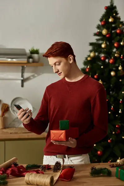 Ein junger Mann im Pullover steht in einer festlichen Küche mit bunt verpackten Geschenken. — Stockfoto