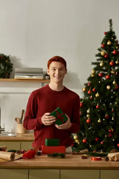 A cheerful young man stands by a decorated kitchen counter, holding a wrapped gift. — Stock Photo