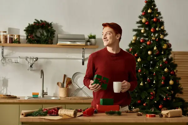 A young man holds a cup and a gift in a decorated kitchen, embracing holiday cheer and warmth. — Stock Photo
