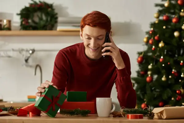 Un joven sonríe mientras habla por teléfono, abriendo con entusiasmo regalos de vacaciones en casa.. - foto de stock