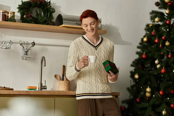 A cheerful young man smiles as he enjoys a cozy drink and holds a Christmas gift in the kitchen. — Stock Photo
