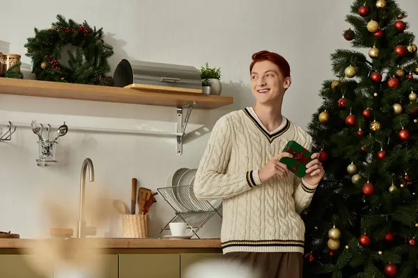 A young man smiles while holding a small gift beside a beautifully adorned Christmas tree. — Stock Photo