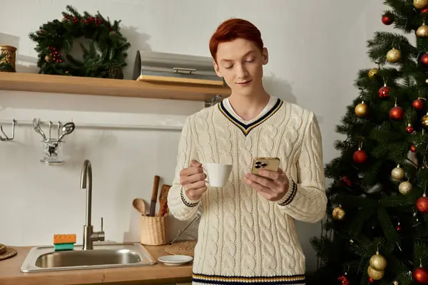 In einer festlichen Küche nippt ein junger Mann an einem heißen Getränk, während er auf seinem Handy blättert. — Stockfoto