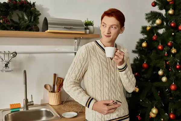 Ein junger Mann hält eine Tasse in der Hand und genießt einen Moment der Freude am Weihnachtsbaum in seiner Küche. — Stockfoto