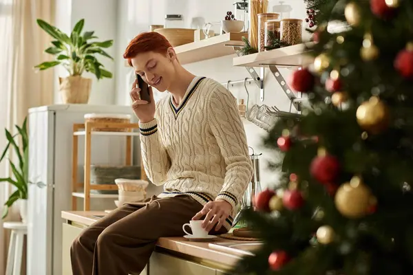 A young man relaxes at home during Christmas, happily chatting and sipping a warm drink. — Stock Photo