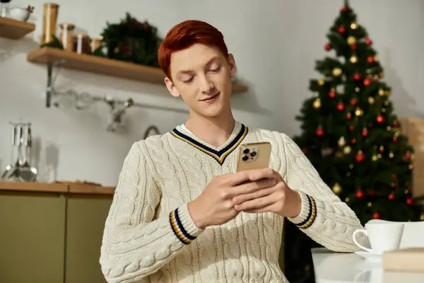 A young man with ginger hair smiles as he relaxes and checks his phone amidst festive decorations. — Stock Photo