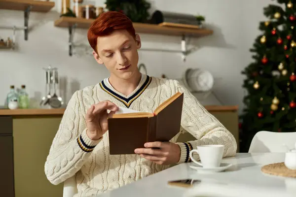 Молодой человек читает книгу за столом, окруженный рождественскими украшениями и ёлкой. — стоковое фото
