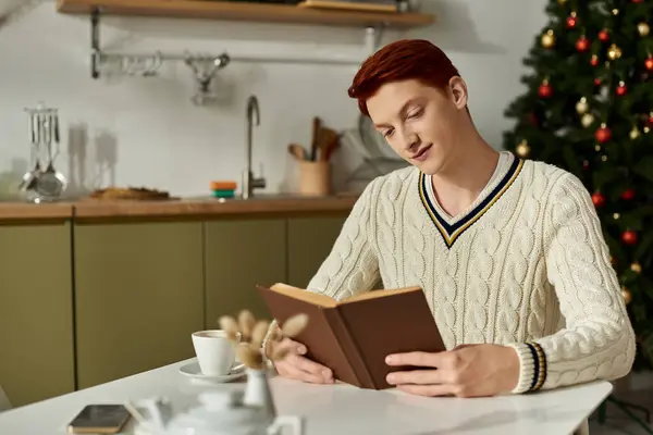A young man sits at a table with a warm cup while immersed in a book on Christmas morning. — Stock Photo