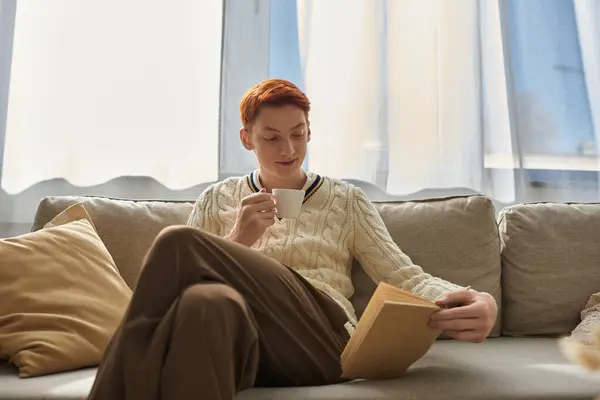 A young man relaxes on a couch, sipping tea while reading a book in a cozy setting. — Stock Photo