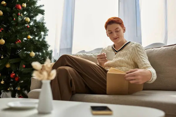 An einem friedlichen Weihnachtsmorgen entspannt sich ein junger Mann mit einem Buch und einem Drink in festlichem Rahmen. — Stockfoto