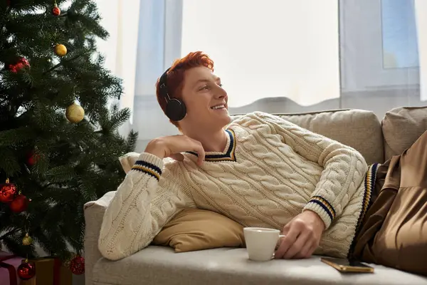 A young man relaxes on the couch, sipping a warm drink and enjoying music this Christmas. — Stock Photo