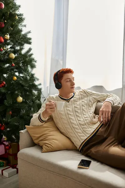 A young man lounges on a couch, sipping a warm drink while listening to music with headphones. — Stock Photo
