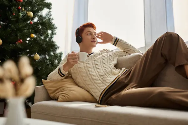 A young man nestled comfortably on the couch, sipping a warm drink while immersed in holiday joy. — Stock Photo