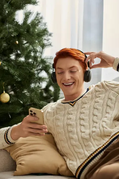 Un giovane si siede comodamente vicino ad un albero di Natale, sorridendo ascoltando musica. — Foto stock