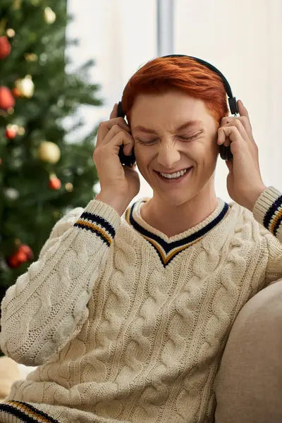 A delighted young man listens to music while seated beside a well decorated Christmas tree. — Stock Photo