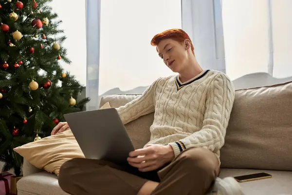 A young man relaxes on the couch, using his laptop in a cozy Christmas setting. — Stock Photo