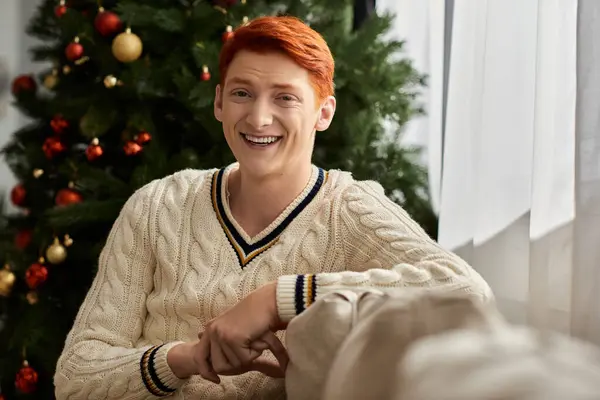 A young man with bright red hair smiles warmly while surrounded by Christmas decor. — Stock Photo