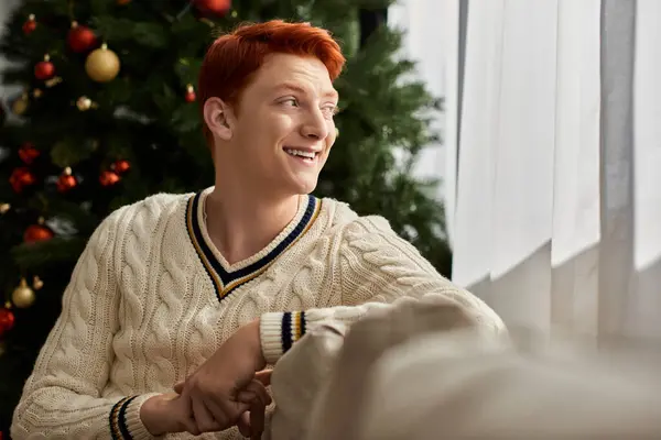 Der junge Mann strahlt vor Glück, als er neben einem Weihnachtsbaum sitzt, der mit Ornamenten gefüllt ist.. — Stockfoto