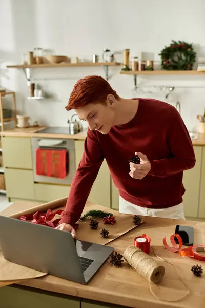 The young man is busy decorating for Christmas while using a laptop in his kitchen. — Stock Photo