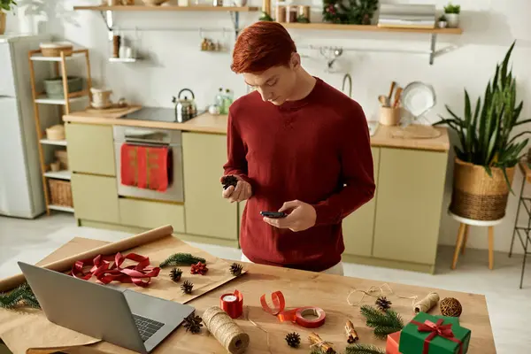 Un joven con un suéter rojo prepara regalos de Navidad en una isla de cocina decorada. - foto de stock