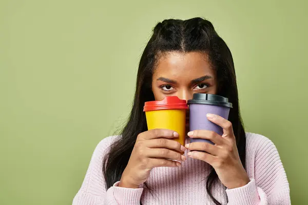 Wearing stylish warm clothing, a young woman playfully holds two colorful cups close to her face. — Stock Photo