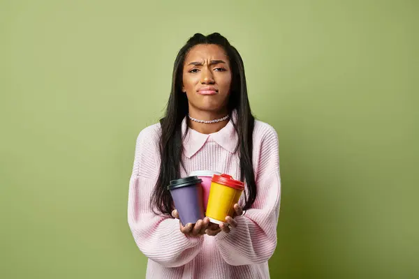 A stylish young woman in warm autumn attire stands against a green backdrop with two coffee cups. — Stock Photo