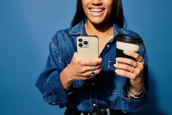 Una mujer alegre con ropa cálida de otoño sonríe, sosteniendo el café y su teléfono frente a una pared azul. - foto de stock