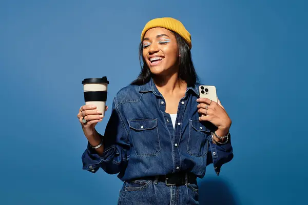 Una giovane donna in caldo abbigliamento autunnale sorride mentre tiene il caffè e uno smartphone contro il blu. — Foto stock