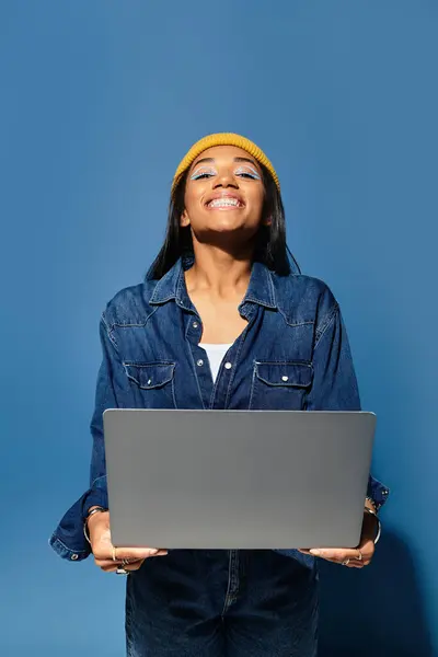 Smiling young woman in a cozy outfit stands with a laptop against a blue background — Stock Photo
