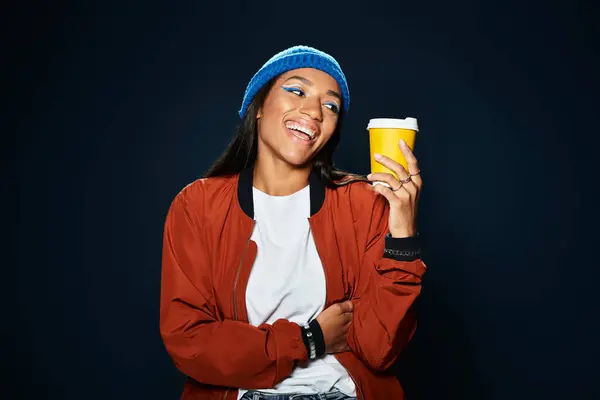 A young woman wears trendy fall attire and holds a yellow cup, exuding joy in a cozy atmosphere. — Stock Photo