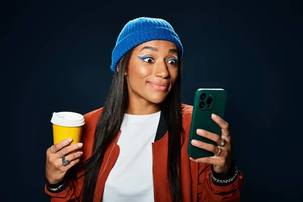 A stylish young woman smiles as she holds her drink and looks at her phone outdoors in autumn. — Stock Photo