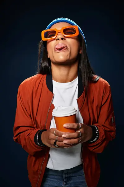 A young woman in cozy autumn fashion enjoys a hot drink while playfully sticking out her tongue. — Stock Photo
