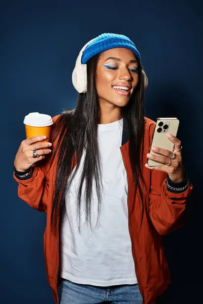 Dressed in warm layers, a stylish young woman smiles while scrolling her phone and sipping coffee. — Stock Photo