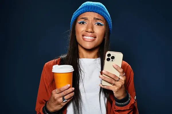A young woman dressed in autumn fashion expresses mixed emotions while holding a drink and phone. — Stock Photo