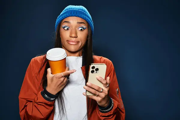 A stylish young woman in warm autumn clothing holds coffee and looks surprised at her phone. — Stock Photo
