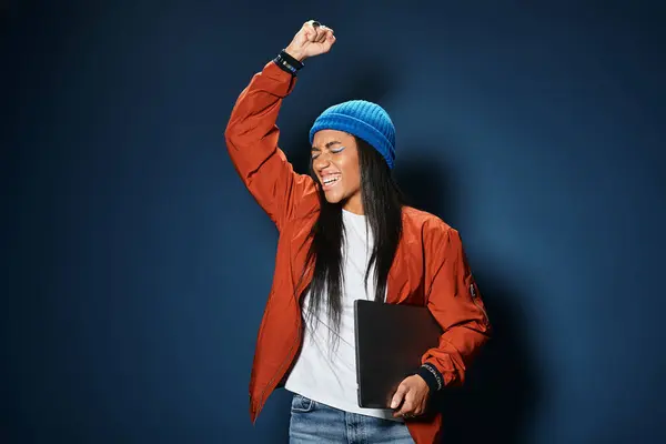 A young woman in a stylish orange jacket and blue beanie raises her fist in exhilaration. — Stock Photo