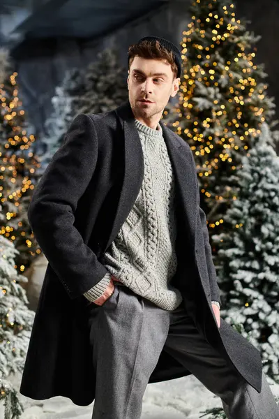 A handsome man dressed in winter attire stands confidently near snow-laden pine trees, capturing holiday spirit. — Stock Photo