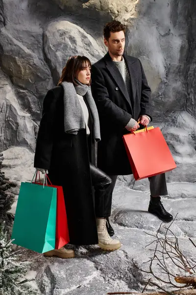 A couple in winter coats stands by snow-laden pine trees, sharing festive spirit with shopping bags. — Stock Photo