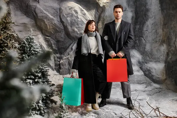A stylish couple embraces the winter spirit, surrounded by snow-covered pine trees — Stock Photo