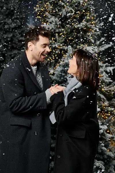 A cheerful couple in winter coats shares a warm moment near snow-covered pine trees during Christmas. — Stock Photo