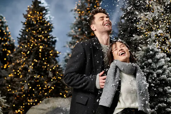 A couple in winter attire laughs joyfully together as snowflakes fall among the illuminated pine trees. — Stock Photo