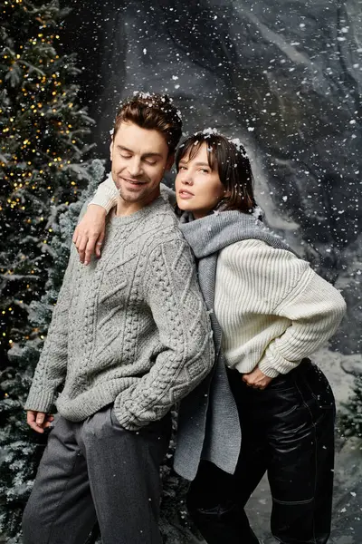 A lovely couple in warm grey sweaters enjoys a festive moment near snow-covered fir trees during winter celebrations. — Stock Photo