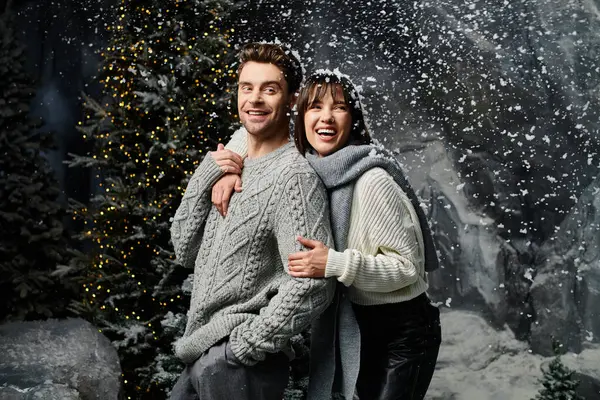 A couple bundled in cozy grey sweaters enjoys a festive winter moment near snow-covered fir trees. — Stock Photo