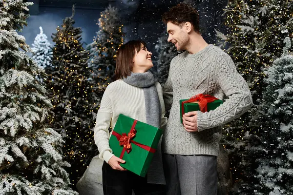 A happy couple shares love and laughter while holding Christmas presents surrounded by snowy fir trees. — Stock Photo