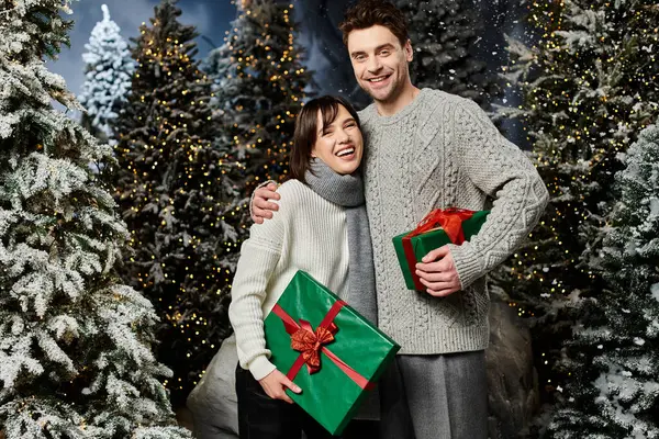 A couple joyfully embraces by snow-covered trees, sharing festive gifts and warm wishes for the holidays. — Stock Photo