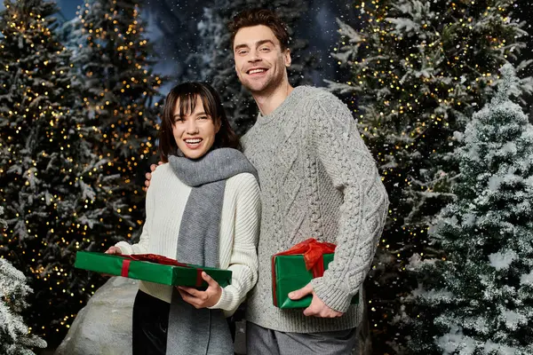 A couple wrapped in warm sweaters smiles happily while exchanging gifts near festive, snow-draped fir trees. — Stock Photo