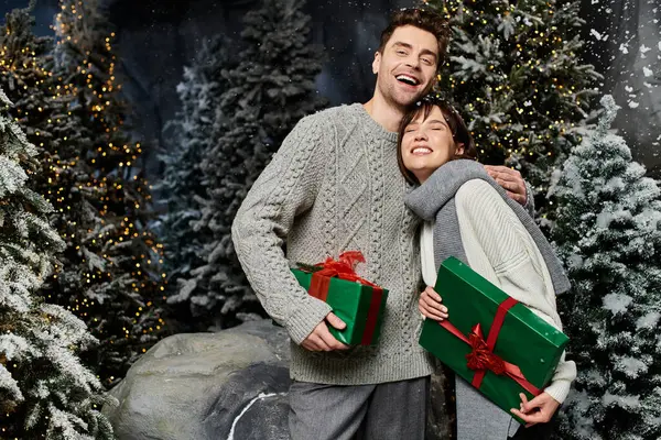 A couple beams with happiness while holding gifts, surrounded by snow-covered fir trees in winter. — Stock Photo