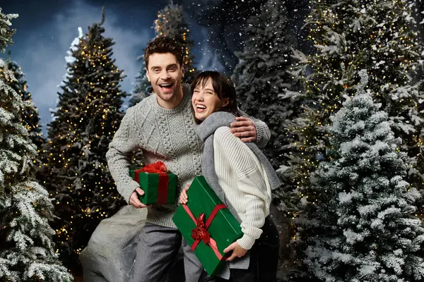 A happy couple embraces the holiday spirit, surrounded by snow-covered trees while holding gifts. — Stock Photo