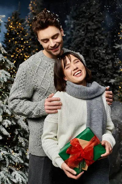 Una coppia felice in maglioni grigi caldi si erge tra abeti innevati, condividendo allegria e amore festivo. — Foto stock