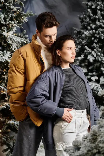 A beautiful couple stands close together, dressed warmly, amidst snow-laden pine trees in winter style. — Stock Photo